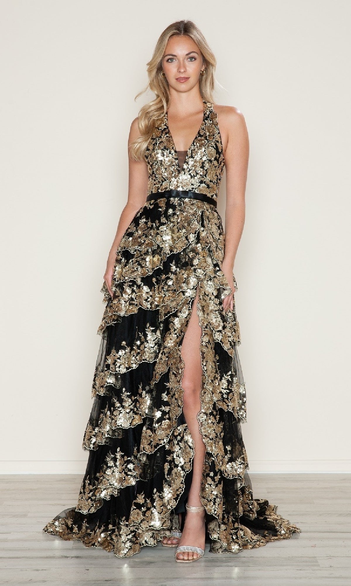 A woman wears the Poly USA 9618 Long Prom Dress, featuring a sleeveless A-line design with gold floral embroidery, plunging neckline, tiered ruffles, high slit, and paired with silver heels.